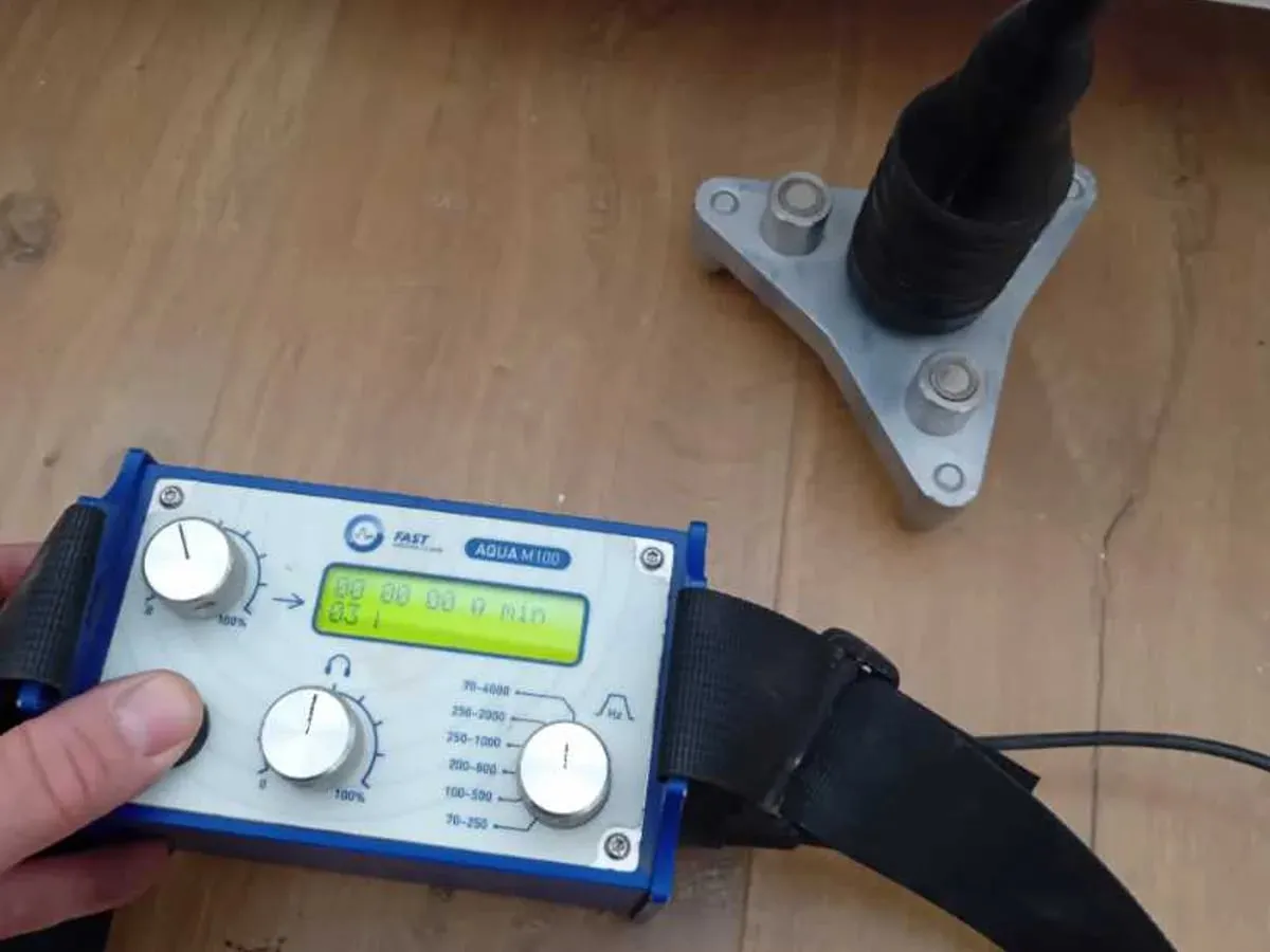 A hand holds a FAST AQUA M100 amplifier while a microphone on a three-legged metal contact foot sits on a bare plywood subfloor