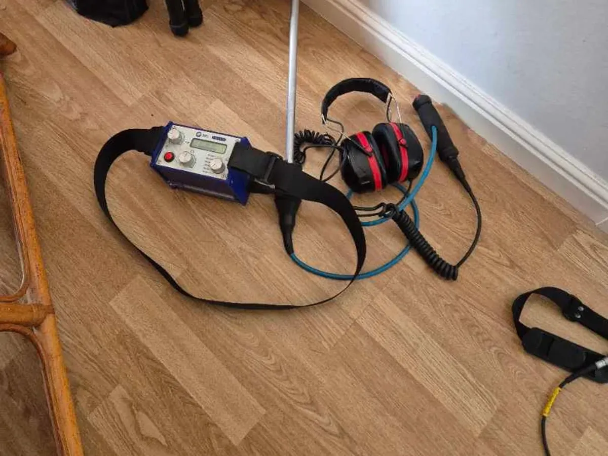 A listening stick, a blue amplifier with a shoulder strap and red ear-defender headphones laid out on a wood-effect floor