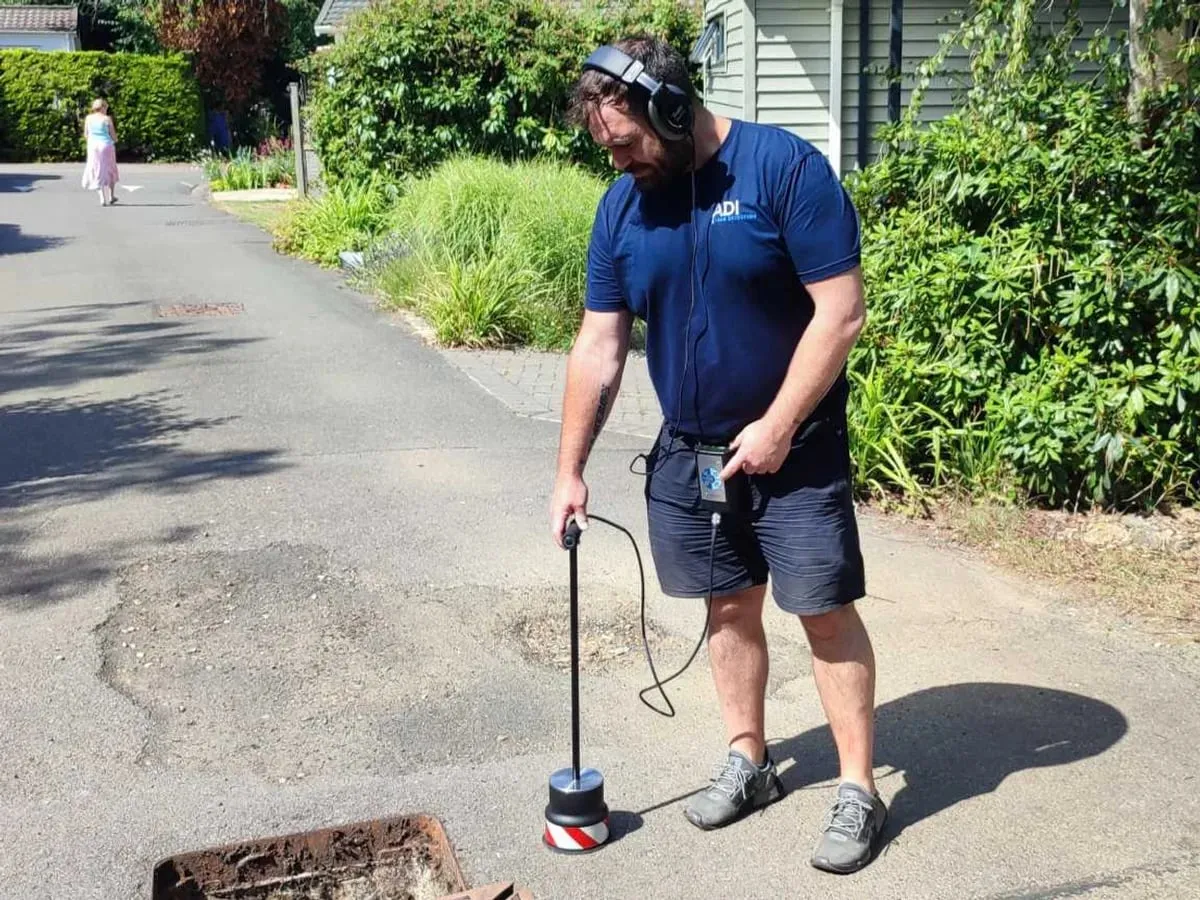 ADI engineer wearing headphones, holding a ground microphone on a pole on a site road beside an open drain cover